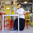 "A woman sitting in a retro-style café, wearing YUI Tabi Shoes in denim, paired with a white graphic T-shirt and a navy skirt, holding a yellow cup and smiling."