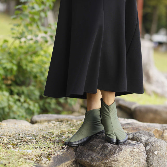 "Close-up of a person wearing U-ten x FLEECE Tabi Shoes in khaki, paired with a black midi skirt, standing on natural stone in an outdoor setting."