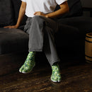 A person sitting on a dark sofa wearing green DISCO Tabi Shoes with a vivid geometric pattern, paired with gray herringbone pants and a white T-shirt.