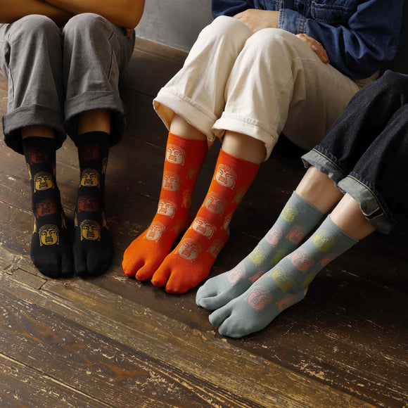 Three people sitting side by side wearing long Tabi Socks 'Nara Daibutsu' in black, orange, and dull blue — each featuring colorful Buddha face designs