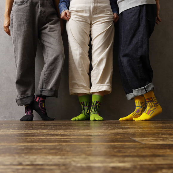 "Three people standing side by side wearing Tabi Socks 'Sankuzushi' in black, green, and yellow, with cuffed pants revealing the colorful designs"