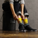 Person wearing yellow Tabi socks “Sankuzushi” with black Tabi shoes “RAIN,” adjusting the socks while sitting on a metal container.