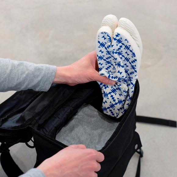 Packing view of blue Tabi Shoes "TIGRE" being placed into a black backpack, showing the patterned soles.